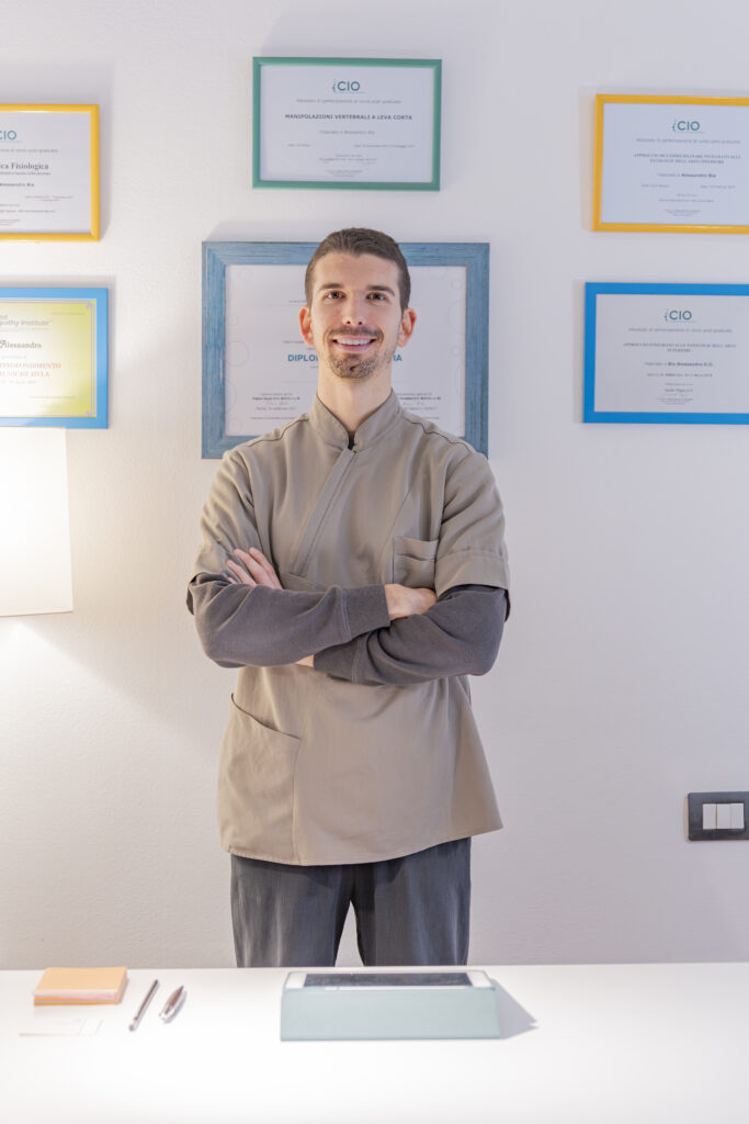 Salute nelle tue mani, il nuovo progetto del CIO Alessandro Bia, osteopata, sorridente in divisa beige con braccia incrociate, davanti a certificati incorniciati appesi al muro.